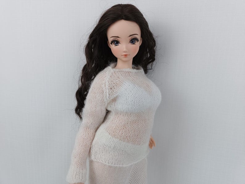 May include: A doll with long, dark brown wavy hair, dressed in a sheer, off-white knit sweater and skirt. The doll has a fair complexion, with detailed facial features and blue eyes. The outfit appears to be handmade.