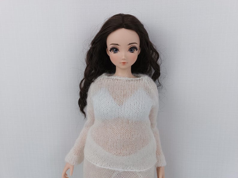 May include: A doll with long, dark brown wavy hair and blue eyes. The doll is wearing a sheer, white knit sweater and a matching skirt. The doll's skin is pale, and it has a neutral expression.