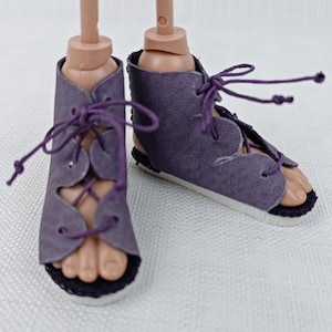 May include: A pair of purple doll sandals featuring a high-top design with scalloped edges and matching purple laces that crisscross through eyelets. The sandals have a flat white sole with a thin black trim along the bottom edge of the footwear.