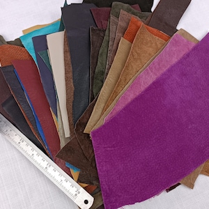 May include: A collection of leather scraps in various colors, including purple, brown, black, and blue. A metal ruler is placed on the left side of the leather pieces. The leather pieces have different textures and finishes.