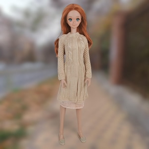 May include: A doll wearing a beige knitted cardigan with a cable knit pattern. The doll has long red hair and is wearing a beige skirt.