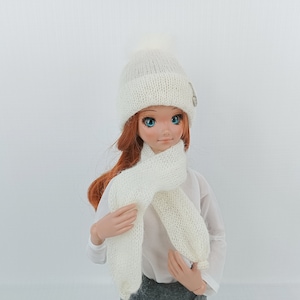 May include: A white knitted hat with a pom-pom on top and a white knitted scarf. The hat has a button on the side.