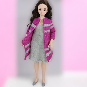 May include: A doll wearing a gray dress and a long, magenta cardigan with gray stripes. The doll has long, dark hair and is standing in front of a pink background. The cardigan is open, revealing the dress underneath.