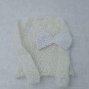 May include: A cream-coloured, knitted sweater with long sleeves and a matching white bow. The sweater is made of a soft, textured yarn. The bow is placed on the front of the sweater. The items are displayed on a white surface.