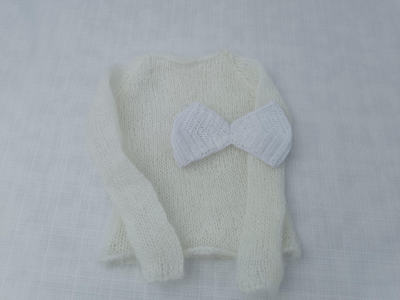 May include: A cream-coloured, knitted sweater with long sleeves and a matching white bow. The sweater is made of a soft, textured yarn. The bow is placed on the front of the sweater. The items are displayed on a white surface.