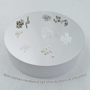 Könnte beinhalten: Eine Sammlung von Puppenkleidungs-Hardware, einschließlich Mini-Knöpfen und Verschlüssen, angeordnet auf einer weißen, kreisförmigen Plattform. Die Hardware umfasst Knöpfe und Verschlüsse in verschiedenen Größen und Stilen in transparenten, weißen und silbernen Farbtönen.