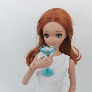 Könnte beinhalten: Eine Puppe mit langen roten Haaren hält ein blaues Glas Martini-Glas.