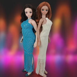 May include: Two fashion dolls stand back-to-back, each wearing a handmade crochet gown. One doll wears a turquoise halter dress with fringe, the other a cream-colored halter dress with fringe. Both dolls have long hair and are posed against a blurred background.