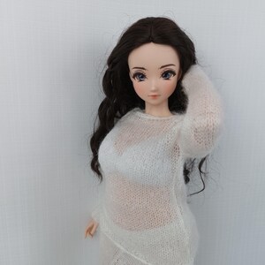 May include: A doll with long, dark hair wearing a sheer white sweater and matching skirt. The doll has a fair complexion and is posed with one arm raised. The background is a plain white wall.