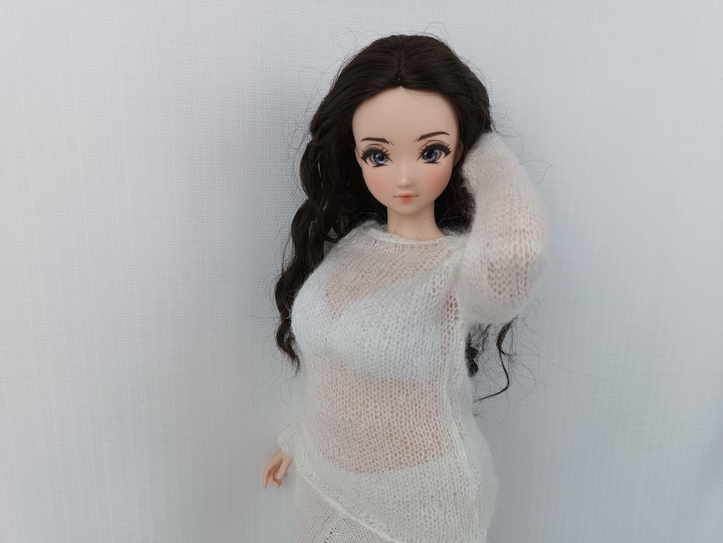 May include: A doll with long, dark hair wearing a sheer white sweater and matching skirt. The doll has a fair complexion and is posed with one arm raised. The background is a plain white wall.
