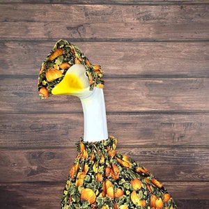 May include: A goose figurine wearing a pumpkin-themed outfit. The outfit includes a bonnet and a dress, both featuring a pattern of orange pumpkins and green leaves on a black background. The goose has a yellow beak and is standing on a green base.