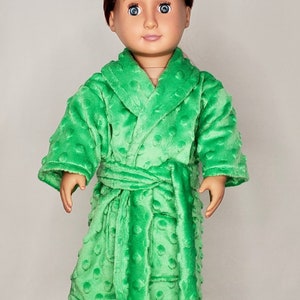 May include: A green plush robe with a tie belt and a hood. The robe is made of a soft, textured fabric. The doll is wearing the robe and white slippers with a dog face design.
