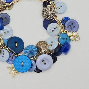 May include: A handmade bracelet featuring a variety of blue buttons in different shades and sizes, accented with gold-tone metal charms. The buttons range from light blue to navy, with some having intricate designs. A star and a cluster of crystals add sparkle.