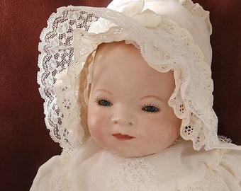 May include: A porcelain baby doll wearing a white lace dress and bonnet. The doll has blue eyes and a painted smile.