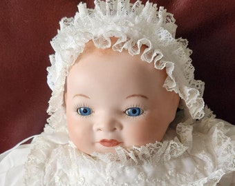 Op de afbeelding: Een porseleinen babypop die een wit kant-doopjurk en mutsje draagt. De pop heeft blauwe ogen en een geschilderde glimlach.