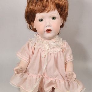 May include: A porcelain doll with red hair wearing a pink dress with lace trim and white shoes.