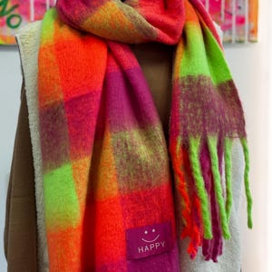 Könnte beinhalten: Ein lebendiger, übergroßer Schal mit Karomuster in Orange-, Rosa-, Grün- und Lilatönen. Der Schal hat einen Fransenrand und ein kleines rechteckiges Etikett mit der Aufschrift "HAPPY" und einem lächelnden Gesicht.