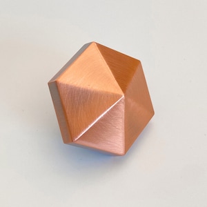 Könnte beinhalten: Ein kupferfarbener Schrankknauf mit einem geometrischen, facettierten Design. Der Knauf hat eine warme, metallische Oberfläche und eine moderne Ästhetik. Die Form ist ein abgestumpftes Rhombikuboktaeder, eine dreidimensionale Form mit 26 Flächen.