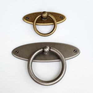 May include: Two oval-shaped cabinet pulls with ring handles. The top pull is gold with a brass finish. The bottom pull is silver with a brushed nickel finish.