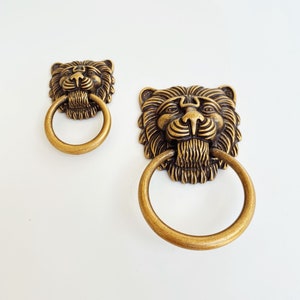 May include: Two antique gold lion head door knobs with ring pulls. The lion heads are detailed with a textured finish.
