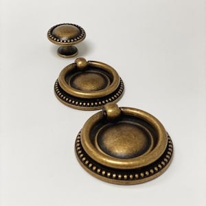 May include: Three antique gold cabinet knobs with a beaded edge. The knobs are round and have a ring pull. The knobs are arranged in a row from smallest to largest.