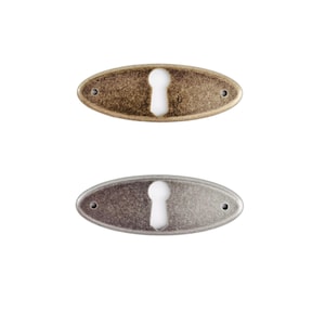 May include: Two oval keyhole escutcheons. One is bronze-toned, the other is silver-toned. Each has a keyhole cutout and two small screw holes. These decorative hardware pieces are for furniture or doors.