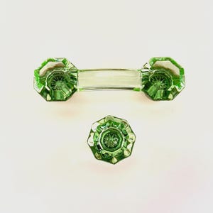 May include: A set of three green glass cabinet hardware pieces. The set includes a rectangular handle with octagonal ends and a single octagonal knob. The glass has a faceted design, set against a white background.