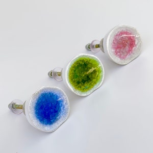 May include: Three decorative cabinet knobs with a geode design. Each knob features a white ceramic base with a colorful, crystal-like center in blue, green, and pink. The knobs have silver-toned hardware for easy installation.