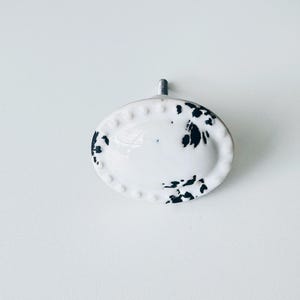 May include: White oval cabinet knob with black speckles and a silver screw. The knob has a raised edge.