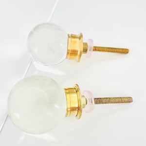 May include: Two clear glass cabinet knobs with gold metal bases and pink plastic washers. The knobs have a round shape and are attached to screws.
