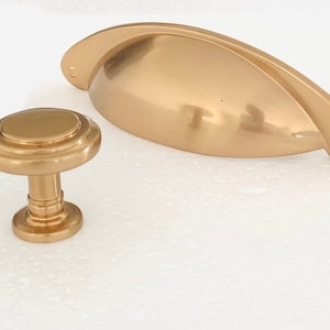 Könnte beinhalten: Zwei goldene Schrankknöpfe, einer ist ein runder Knopf mit erhöhtem Rand und der andere ist ein gebogener, ovaler Griff.