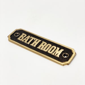 Puede incluir: Un letrero de metal dorado con fondo negro y las palabras "BATH ROOM" en letras doradas.
