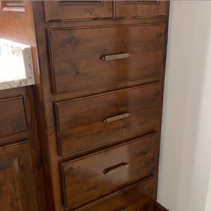 May include: A set of five wooden drawers with brushed gold hardware. The drawers are in a bathroom vanity and are made of a dark wood.