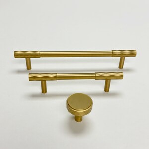 May include: Three gold cabinet hardware pulls with a geometric pattern. Two are bar pulls and one is a round knob.