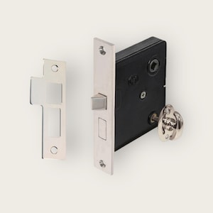 Pode incluir: Uma fechadura de porta prateada com uma trava preta e uma maçaneta prateada. A fechadura tem formato quadrado e está montada em um fundo branco.