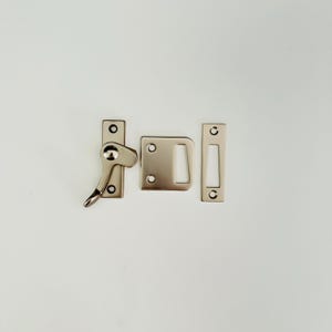 May include: A silver window latch with a lever handle and two strike plates. The latch is designed to secure a window in the open position.