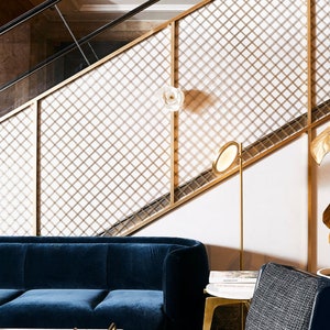 May include: A blue velvet sofa sits in front of a gold metal grid wall. A gold floor lamp with a round white shade is next to the sofa. A book titled "Remarkable Hospitality Design" is on a small table next to the sofa.