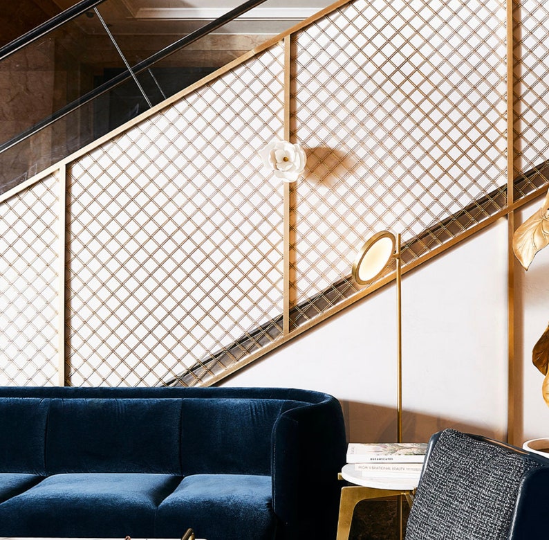 May include: A blue velvet sofa sits in front of a gold metal grid wall. A gold floor lamp with a round white shade is next to the sofa. A book titled "Remarkable Hospitality Design" is on a small table next to the sofa.