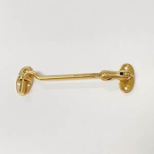 May include: A gold-toned metal door hook with a curved arm and two round plates. The hook is designed to be mounted on a door or window to secure it.