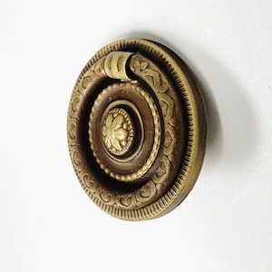 Puede incluir: Un tirador de armario redondo y adornado de latón con un asa de anillo decorativa. El tirador tiene un acabado marrón oscuro con detalles dorados.