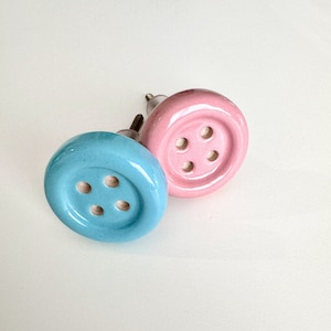 May include: Two ceramic cabinet knobs shaped like buttons. One is light blue with four holes and the other is pink with four holes.