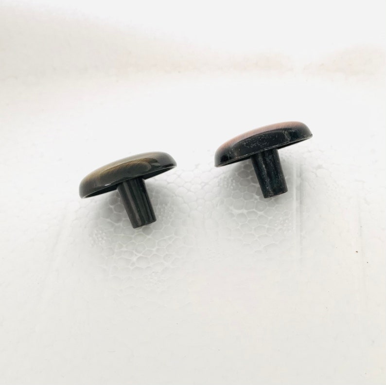 May include: Two small, round, black and brown plastic knobs with a short, black stem. The knobs are on a white surface.