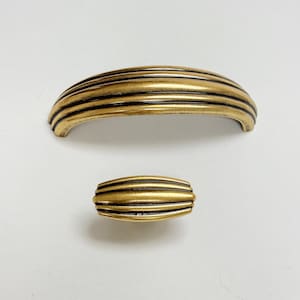 Könnte beinhalten: Ein Satz aus zwei goldfarbenen Schrankknöpfen mit geripptem Design. Der größere Knopf ist gewölbt und der kleinere Knopf ist oval.