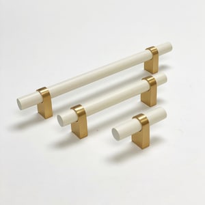 Op de afbeelding: Vier witte kastgrepen met gouden metalen bases. De grepen zijn in verschillende maten, waarbij de grootste ongeveer 25 cm lang is en de kleinste ongeveer 7,5 cm lang.