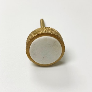 May include: A round, gold-colored knob with a white marble center. The knob has a textured edge and a screw on the back.