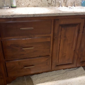 May include: A wooden bathroom vanity with three drawers and two cabinet doors. The vanity has a light brown stain and gold-colored hardware.