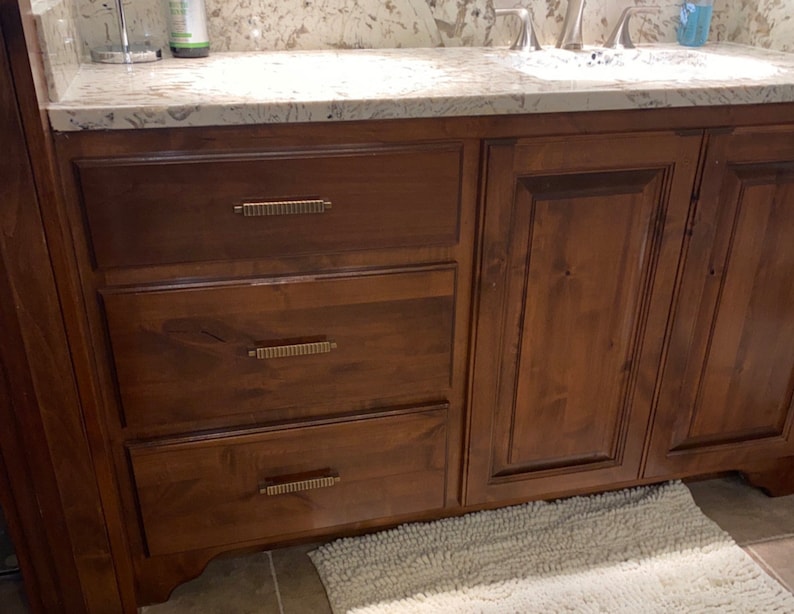 May include: A wooden bathroom vanity with three drawers and two cabinet doors. The vanity has a light brown stain and gold-colored hardware.