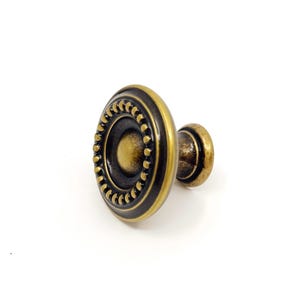 May include: A round cabinet knob with an antique brass finish, featuring a central dome surrounded by a ring of small raised beads and a dark recessed border. The hardware has a stepped profile and a cylindrical base for mounting to furniture.