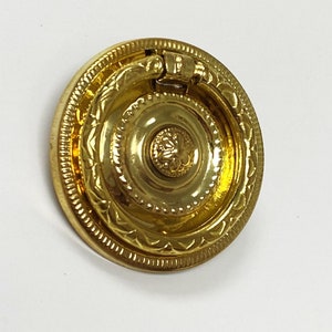 May include: Gold-toned metal ring pull cabinet knob with a decorative, ornate design. The knob has a round base with a raised ring and a smaller, centered ring.