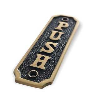 Op de afbeelding: Een messing duwschild met een zwarte, getextureerde achtergrond. Het bord heeft de tekst "PUSH" in verticale gouden letters.
